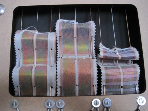These flexible solar cells are coated directly onto woven polyester fabric, using a sandwich configuration of conducting layers and a photo-active thin film of silicon. The fabric strips are mounted in a test jig with guide wires so that they may be bent and stretched repeatedly by the central cords to demonstrate their flexibility while delivering electrical power. Colors change according to the illumination direction. Photo: Power Textiles Ltd.